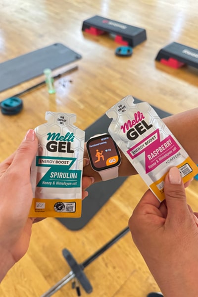 Two hands holding Melli Gel packets with a blurred background of exercise equipment.